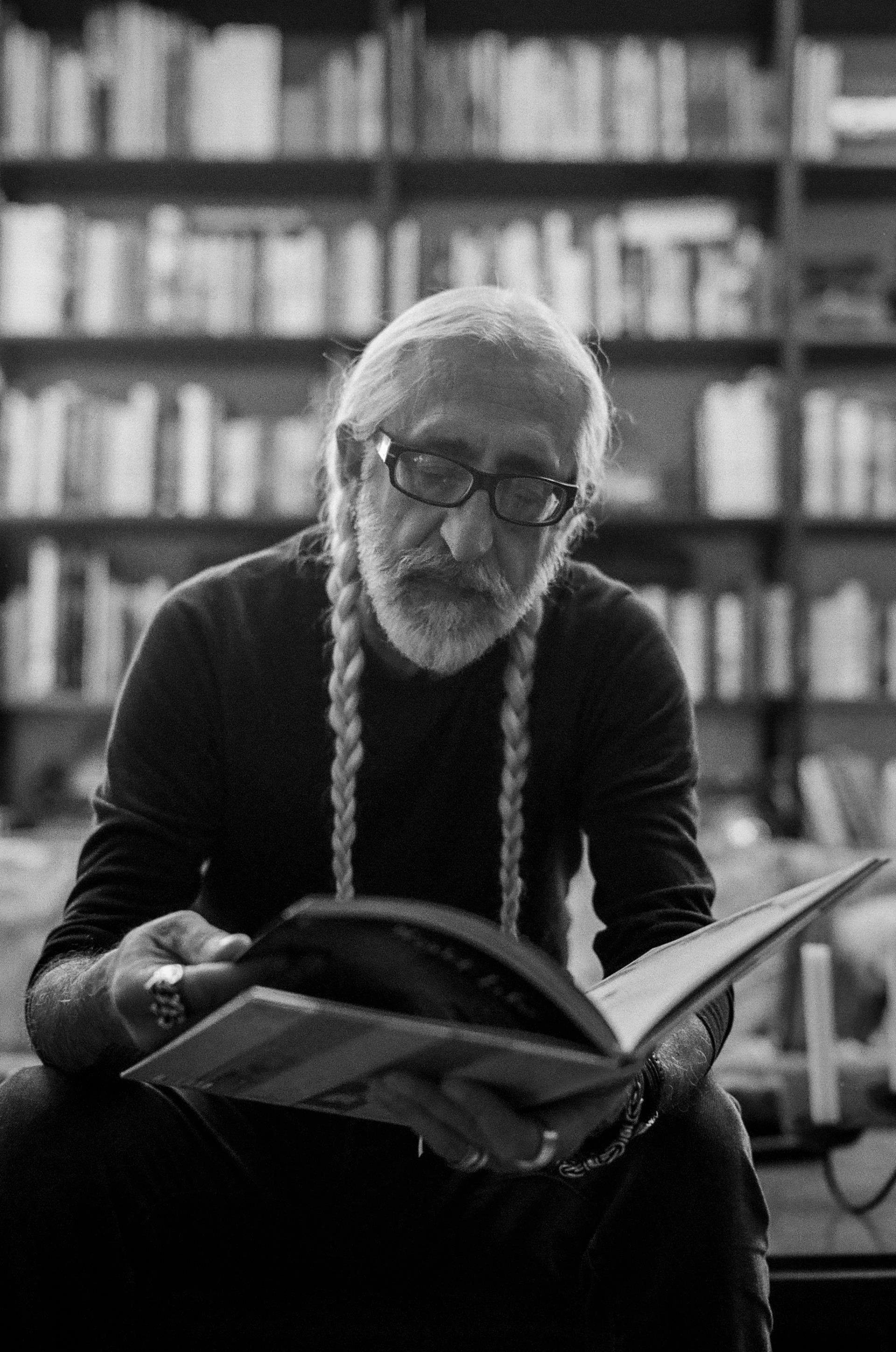 Vahe Berberian black and white photograph. He has white hair, wears glasses, has a beard, and is reading a book with a bookshelf in the background.